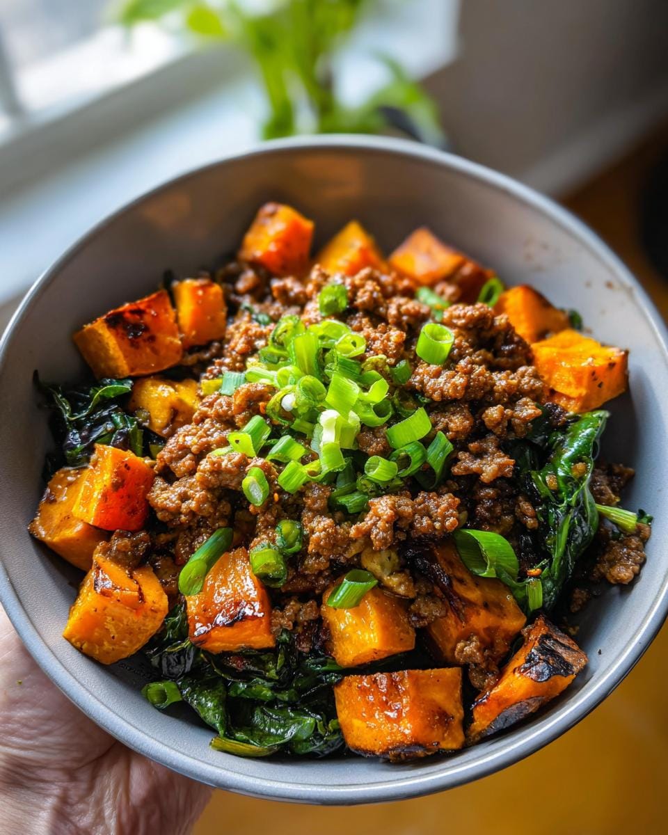A close-up of a sweet potato ground beef protein bowl, featuring roasted sweet potato cubes, seasoned ground beef, and wilted greens, topped with chopped scallions.