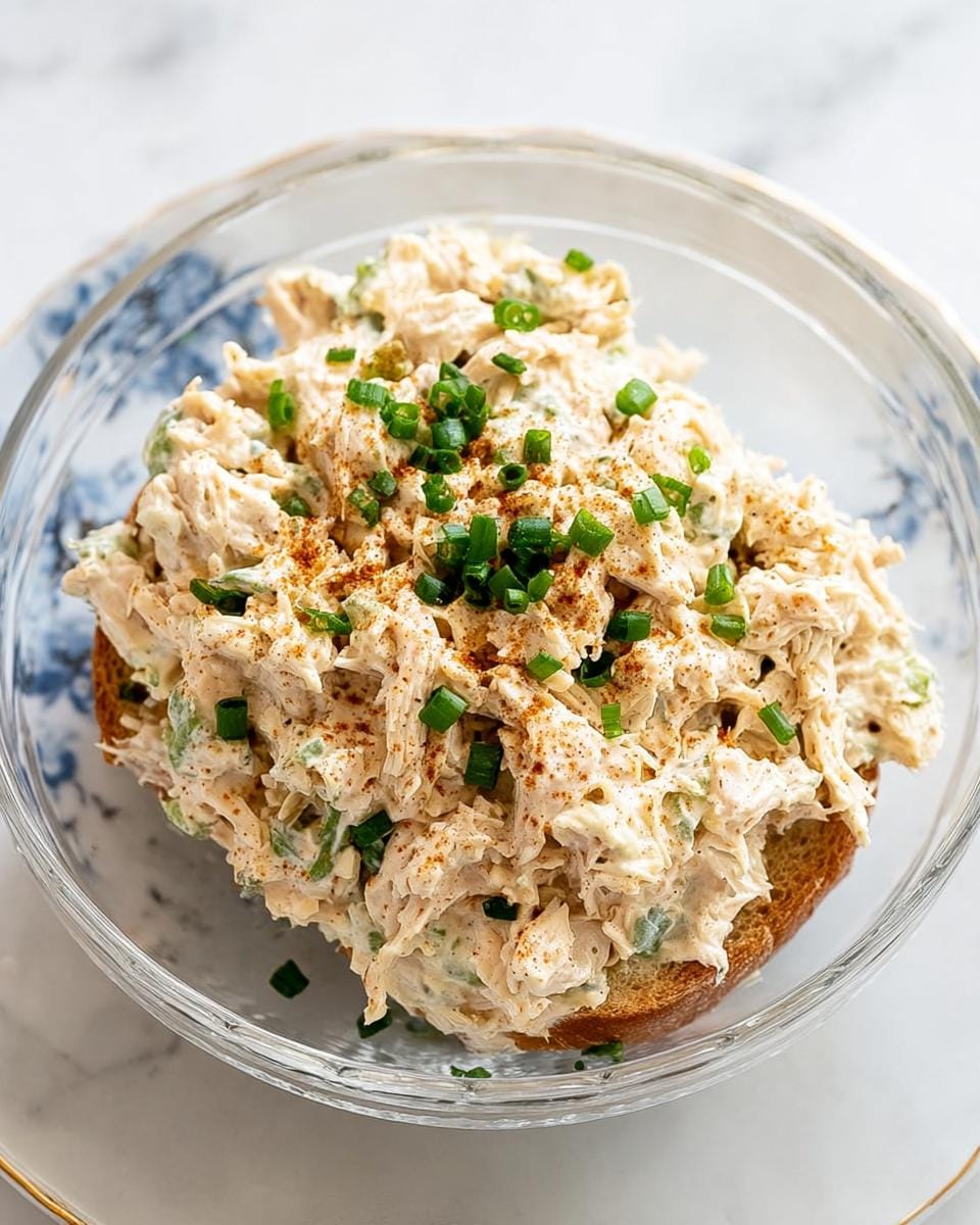 Close-up of a generous serving of creamy chicken salad piled high on a slice of toasted bread, garnished with chives and paprika.