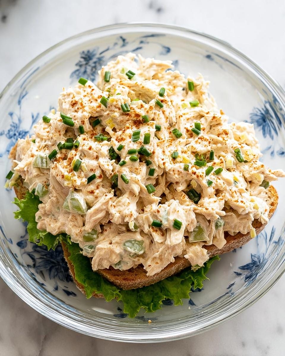 A generous portion of creamy chicken salad piled high on whole wheat bread with lettuce, garnished with chives and paprika.